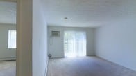 This single-floor space offers a clean, straightforward layout designed for comfortable everyday living. The main living area is finished with wall-to-wall carpeting that lends a soft, quiet feel underfoot, while the textured ceiling adds a subtle layer of visual character to the room.

A prominent sliding glass door serves as the focal point of the living area, fitted with vertical blinds that allow for flexible light control. The door opens to an outdoor space beyond, where surrounding greenery is visible, bringing a sense of the outdoors in and flooding the room with natural light.

Practical climate control is provided by a wall-mounted air conditioning unit, conveniently positioned near the sliding door for efficient airflow throughout the space. The walls are finished in a clean, neutral white, creating a bright and open atmosphere that serves as a versatile backdrop for a variety of interior arrangements.

Overall, the space presents a simple yet functional environment with ample natural light, neutral finishes, and essential amenities that make it a practical and adaptable living area.