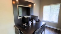 This single-floor space is thoughtfully designed as a functional business center, offering a sleek and professional environment suited for work and productivity. The focal point of the room is a large wall-mounted flat-screen display set against a bold, dark accent wall, creating a striking visual backdrop that adds depth and sophistication to the space. Below the display, a spacious dark-finish work desk is equipped with multiple computer monitors, a keyboard, mouse, and readily available resource materials, making it a fully operational workstation. Two patterned upholstered chairs with a contemporary design flank the desk, providing comfortable seating for users. Pendant lighting with an industrial-inspired aesthetic hangs from the ceiling, casting warm, focused light across the workspace. A window fitted with horizontal blinds allows natural light to filter into the room, contributing to a bright and inviting atmosphere while maintaining privacy. The dark hardwood-style flooring complements the overall color palette of deep neutrals and grays, lending a polished and cohesive look throughout. The space balances form and function, making it well-suited as a shared business resource area.