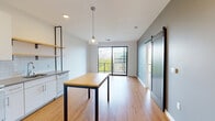 This contemporary 2-bedroom, 1-bathroom unit offers a well-designed, efficient layout spread across a single floor. The open-concept kitchen and living area serves as the heart of the home, featuring light hardwood-style flooring, recessed lighting, and a stylish pendant light above a central dining table. The kitchen is equipped with white cabinetry, a white subway tile backsplash, stainless steel refrigerator, and open wooden shelving mounted on black industrial-style brackets. A sliding barn door with frosted panel detailing adds a modern design accent to the living space. Floor-to-ceiling sliding glass doors lead to a private balcony, flooding the living area with natural light and offering elevated views of the surrounding cityscape and greenery. The primary bedroom features plush carpeting, large windows with city views, and access to a secondary balcony. The second bedroom is equally bright, with soft carpet and a bold dark wood door. The full bathroom includes a white vanity with gray countertop, a large mirror, and tile flooring. A utility room completes the unit. Clean white walls and a neutral palette throughout create a bright, cohesive, and modern atmosphere.