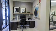This single-floor business center space is thoughtfully designed to provide a functional and visually refined environment for focused work. The centerpiece of the room is a built-in workstation featuring a granite-topped desk with dark cabinetry beneath, offering ample storage and a sleek, contemporary look. A black ergonomic office chair is positioned at the workstation, which is equipped with a desktop computer monitor and a printer, making it a fully operational workspace. Above the desk, a floating wood shelf adds a warm contrast to the textured gray wallcovering that serves as a striking accent feature. The shelf is styled with framed black-and-white artwork, a small potted plant, and decorative objects, lending the space a curated, sophisticated feel. A large abstract painting in blue, gold, and black tones adorns the adjacent white wall, adding a bold artistic element to the room. Light-toned tile flooring ties the space together, reflecting light and contributing to the bright, clean ambiance. Crown molding along the ceiling adds a finishing architectural touch. Overall, the space balances professional utility with modern design sensibility.