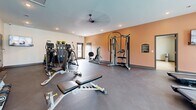 Located at 411 Sage Rd N in White House, Tennessee, this fitness center offers a comprehensive, single-level layout thoughtfully divided into two distinct functional areas. The main workout room features durable rubber flooring and is outfitted with a broad selection of strength and cardio equipment, including treadmills, ellipticals, a cable machine, weight benches, and a full dumbbell rack ranging up to 50 lbs. Large wall-mounted mirrors amplify the sense of space and allow users to monitor their form. Recessed lighting and a ceiling fan keep the environment bright and comfortable, while windows provide views of the surrounding community and pool area. Warm accent walls and tasteful greenery add an inviting atmosphere to the workout floor. The second area serves as a dedicated studio space, finished with warm hardwood-style flooring and equipped for functional and group fitness training. This room features wall-mounted resistance bands, stability balls, kettlebells, a TRX suspension system, and a full floor-to-ceiling mirror along one wall. Multiple windows flood the studio with natural light, making it an ideal setting for yoga, stretching, or guided fitness classes. Together, both spaces form a well-rounded fitness facility suited for a variety of training styles and fitness levels.