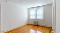 This single-floor space presents a clean, functional layout across several rooms, each finished with warm-toned hardwood flooring that reflects natural light throughout. The main living area is open and well-lit, featuring a large window fitted with blinds and a classic cast-iron radiator beneath it, providing an efficient and traditional heating solution. White walls and a drop-panel ceiling contribute to a bright, neutral ambiance throughout the space.

The kitchen is compact yet practical, equipped with dark wood-toned cabinetry — both upper and lower — offering ample storage. A stainless steel sink is set into a light-colored countertop, and a white gas range with overhead ventilation completes the cooking area. The floor transitions to tile in this space, providing a durable and easy-to-clean surface.

The bathroom includes a full-size bathtub with a tub/shower combination, outfitted with chrome fixtures and white tile surround, maintaining the clean, minimal aesthetic seen throughout the unit.

The floor plan, visible in the dollhouse view, reveals a straightforward and efficient arrangement of rooms, with clear sightlines between spaces and easy accessibility. Overall, this space is characterized by its classic finishes, natural light, and practical design.