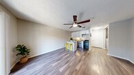 This single-floor unit offers a thoughtfully designed open-concept layout that seamlessly connects the living area and kitchen. Rich wood-look flooring runs throughout the space, complemented by neutral-toned walls and a textured ceiling. A ceiling fan with an integrated light fixture anchors the main living area, which is generously sized and filled with natural light from a sliding glass door that opens to a private balcony overlooking a landscaped outdoor setting.

The kitchen is efficiently designed and features white shaker-style cabinetry, granite-look countertops, a white subway tile backsplash, a stainless steel sink, a dishwasher, and a black range with an overhead exhaust hood. A peninsula with seating for two on metal bar stools creates a casual dining and gathering space that bridges the kitchen and living room.

A hallway leads from the main living area to the bathroom, which is finished with matching wood-look flooring, white cabinetry with granite-look countertops, and a full tub-and-shower combination with white tile surround. The overall design is clean, modern, and cohesive, making efficient use of the available space while maintaining a bright and welcoming ambiance throughout.