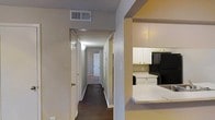 This single-story space is organized around a central hallway that serves as the main artery connecting the various rooms. The corridor extends toward the back of the unit, with multiple doorways branching off on either side, providing a clear and practical flow throughout the interior. The walls feature a lightly textured finish, and the flooring transitions from darker material in the main areas to carpet in the rooms at the end of the hall.

Adjacent to the hallway, the kitchen is visible and features white cabinetry that provides ample storage space. A black refrigerator anchors one end of the kitchen, while a white range or cooktop is positioned nearby. The countertop appears light-colored and extends to form a partial partition between the kitchen and the adjacent area, adding a sense of openness between the spaces.

Overhead HVAC vents are present throughout, ensuring climate control across the layout. Natural light filters in through doorways at the end of the hallway, brightening the interior. The overall design is straightforward and practical, prioritizing functional use of space with a neutral color palette throughout.