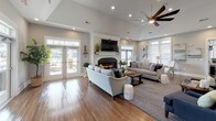 Located on Broad Street in Sumter, South Carolina, this single-floor clubhouse offers a thoughtfully designed community gathering space that blends comfort with refined style. The main lounge is the heart of the facility, boasting soaring vaulted ceilings, warm hardwood-style flooring, and a central fireplace framed by a classic white mantle. Large French doors open directly to an outdoor patio and pool area, flooding the room with natural light and providing seamless indoor-outdoor connectivity. A ceiling fan overhead and recessed lighting complement the bright, airy atmosphere.

The fully equipped kitchen features white shaker cabinetry, granite countertops, a gray subway tile backsplash, and stainless steel appliances. A generous island with bar seating creates a natural gathering point between the kitchen and the open-plan living area. Adjacent to the kitchen, a casual dining space with a round table and upholstered seating offers a relaxed setting for residents.

A separate area provides access to a private office or conference room through glass-paned French doors, complete with a small bistro-style table for informal meetings. Throughout the space, soft gray walls, crown molding, and carefully curated artwork contribute to a cohesive coastal-inspired aesthetic. The overall ambiance is welcoming, polished, and well-suited for community gatherings and everyday resident use.