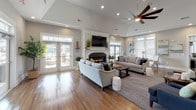 Located on Broad Street in Sumter, South Carolina, this single-floor clubhouse offers a thoughtfully designed community gathering space that blends comfort with refined style. The main lounge is the heart of the facility, boasting soaring vaulted ceilings, warm hardwood-style flooring, and a central fireplace framed by a classic white mantle. Large French doors open directly to an outdoor patio and pool area, flooding the room with natural light and providing seamless indoor-outdoor connectivity. A ceiling fan overhead and recessed lighting complement the bright, airy atmosphere.

The fully equipped kitchen features white shaker cabinetry, granite countertops, a gray subway tile backsplash, and stainless steel appliances. A generous island with bar seating creates a natural gathering point between the kitchen and the open-plan living area. Adjacent to the kitchen, a casual dining space with a round table and upholstered seating offers a relaxed setting for residents.

A separate area provides access to a private office or conference room through glass-paned French doors, complete with a small bistro-style table for informal meetings. Throughout the space, soft gray walls, crown molding, and carefully curated artwork contribute to a cohesive coastal-inspired aesthetic. The overall ambiance is welcoming, polished, and well-suited for community gatherings and everyday resident use.
