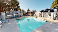 This outdoor amenity space is designed with a resort-inspired aesthetic, centered around a generously sized swimming pool with clear, blue-green water. The pool features a geometric shape with a wide surrounding deck, offering plenty of room for sunbathing and relaxation. Blue lounge chairs line the poolside, providing comfortable spots to unwind under the open sky. On the opposite end, a shaded seating area with red market umbrellas and patio furniture offers a welcoming retreat from the sun. The entire pool area is enclosed by a wrought-iron fence, ensuring privacy and security while maintaining an open, airy feel. Mature trees border the space, adding natural greenery and additional shade. Adjacent to the pool area, a dedicated outdoor kitchen station features dual stainless steel built-in gas grills mounted on a solid concrete base, ideal for outdoor entertaining and communal gatherings. The surrounding residential buildings feature a neutral stucco exterior with pitched rooflines, contributing to the cohesive architectural character of the community. The space benefits from abundant natural light thanks to clear, open skies, making it an inviting and functional outdoor destination year-round.