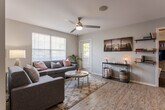 This single-story unit is designed with an open, functional layout that connects the living area and kitchen seamlessly. The living room is bright and welcoming, featuring wood-look flooring, a ceiling fan, large windows that invite natural light, and direct access to an exterior door. The space comfortably accommodates multiple seating areas, making it well-suited for both relaxation and socializing.

The kitchen is positioned adjacent to the living area and features warm wood-toned cabinetry, dark countertops, black appliances including a refrigerator, microwave, and range, as well as a breakfast bar with bar-stool seating. A bar-height counter separates the kitchen from the main living space, enhancing the open feel while maintaining a defined cooking zone.

The bedroom offers carpeted flooring, a window with outdoor views, and a dedicated study nook with a desk and chair — a practical feature for work or study. The room provides a comfortable, quiet retreat with sufficient space for a full bed setup and storage.

Two bathrooms serve the unit, both finished with wood-look flooring. One features a single-sink vanity with warm wood cabinetry and light countertops, while the other offers a clean, simply appointed tub-shower combination. Both are neatly laid out and functional.