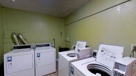 This single-floor shared laundry facility is a practical, no-frills space designed for efficient use. The room is finished with light green painted walls and a textured ceiling, creating a simple and utilitarian environment. The facility is equipped with multiple commercial-grade top-loading washing machines and front-loading dryers, arranged to maximize accessibility and ease of use throughout the room. The machines are coin-operated, catering to a shared-use setting. The flooring consists of neutral tile, which is well-suited for the functional demands of a laundry space. Overhead piping runs along the upper portion of the walls, consistent with the mechanical requirements of the appliances. A utility basket is available within the room for added convenience. The layout is straightforward, allowing users to move between machines with ease. Overall, the space is purpose-built and efficiently organized to meet the day-to-day laundry needs of its users, prioritizing practicality and accessibility above all else.