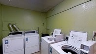 This single-floor shared laundry facility is a practical, no-frills space designed for efficient use. The room is finished with light green painted walls and a textured ceiling, creating a simple and utilitarian environment. The facility is equipped with multiple commercial-grade top-loading washing machines and front-loading dryers, arranged to maximize accessibility and ease of use throughout the room. The machines are coin-operated, catering to a shared-use setting. The flooring consists of neutral tile, which is well-suited for the functional demands of a laundry space. Overhead piping runs along the upper portion of the walls, consistent with the mechanical requirements of the appliances. A utility basket is available within the room for added convenience. The layout is straightforward, allowing users to move between machines with ease. Overall, the space is purpose-built and efficiently organized to meet the day-to-day laundry needs of its users, prioritizing practicality and accessibility above all else.