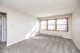 This well-organized single-floor unit offers a practical and comfortable layout across five distinct spaces: a living area, kitchen, bedroom, bathroom, hallway, and closet. The carpeted living room is bright and open, featuring a large multi-pane window with wood-trimmed framing that allows generous natural light to fill the room. A baseboard heating system runs along the exterior wall, and a textured ceiling adds a classic touch to the space. The entryway transitions from hardwood-style flooring into the soft carpet of the main living area. The kitchen is equipped with warm-toned wood cabinetry, a white electric range, a full-size refrigerator, and a double-basin sink beneath under-cabinet lighting. Light wood-style flooring and a tile backsplash complete the functional cooking space. The bedroom is a comfortable size, featuring carpet flooring, a window with natural light, a wall-mounted air conditioning unit, and a baseboard heater. A dedicated closet is accessible directly from the bedroom for convenient storage. The bathroom includes a full tub and shower combination, a wood-vanity with sink, and tiled accents, presenting a clean and classic finish. A hallway connects the rooms efficiently, completing this thoughtfully arranged unit.