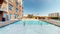 Located in the heart of Charlotte's West Trade Street corridor, this outdoor amenity space offers a well-appointed retreat within a classic multi-story brick residential building. The centerpiece is a generously sized swimming pool with stainless steel entry rails, framed by a spacious brick-paved deck ideal for lounging and socializing. A modern pergola structure with horizontal slat panels provides partial shade and serves as an architectural focal point, while string lights add ambiance for evening use. Adjacent to the pool, a fully equipped outdoor kitchen and grilling station features stone-clad counters and multiple gas grills, making it well-suited for outdoor entertaining. The surrounding deck is outfitted with multiple chaise lounges and high-top bistro seating, offering flexible areas for relaxation. The courtyard area is enclosed by the building's warm brick façade, with multi-level balconies overlooking the space, creating an intimate and private atmosphere. Lush landscaping with seasonal plantings and greenery softens the hardscape throughout. The Charlotte skyline is visible beyond the rooftop-level pergola, providing an urban backdrop. Building access points are conveniently integrated into the courtyard level, connecting residents directly to the amenity area.