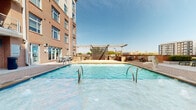 Located in the heart of Charlotte's West Trade Street corridor, this outdoor amenity space offers a well-appointed retreat within a classic multi-story brick residential building. The centerpiece is a generously sized swimming pool with stainless steel entry rails, framed by a spacious brick-paved deck ideal for lounging and socializing. A modern pergola structure with horizontal slat panels provides partial shade and serves as an architectural focal point, while string lights add ambiance for evening use. Adjacent to the pool, a fully equipped outdoor kitchen and grilling station features stone-clad counters and multiple gas grills, making it well-suited for outdoor entertaining. The surrounding deck is outfitted with multiple chaise lounges and high-top bistro seating, offering flexible areas for relaxation. The courtyard area is enclosed by the building's warm brick façade, with multi-level balconies overlooking the space, creating an intimate and private atmosphere. Lush landscaping with seasonal plantings and greenery softens the hardscape throughout. The Charlotte skyline is visible beyond the rooftop-level pergola, providing an urban backdrop. Building access points are conveniently integrated into the courtyard level, connecting residents directly to the amenity area.