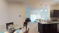 This thoughtfully designed single-floor residence offers a seamless open-concept layout connecting the kitchen, dining, and living areas. The kitchen is equipped with stainless steel appliances, including a refrigerator, range, and microwave, complemented by dark cabinetry, granite-style countertops, a breakfast bar with pendant lighting, and wood-look flooring. The adjacent dining area accommodates a round glass table for four and flows naturally into the carpeted living room, which features a large sliding glass door that floods the space with natural light and provides access to the outdoors. The primary bedroom is generously sized with plush carpeting, a wide window with outdoor views, and direct access to a spacious walk-in closet outfitted with wire shelving. The en-suite bathroom includes a walk-in shower, granite-top vanity, and tile flooring with a natural stone appearance. A second bedroom offers comfortable proportions, carpeted floors, and ample natural light. A second bathroom with a shower and granite vanity serves this bedroom via a connecting hallway. Warm neutral tones throughout the residence create a calm, cohesive ambiance, while consistent finishes and thoughtful layouts provide both comfort and functionality across every room.