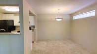 This single-floor space presents a clean, functional layout designed for comfortable everyday living. The main living area is finished with plush, light-beige carpeting that adds warmth underfoot, while the walls are painted in a soft, neutral tone that promotes a calm and airy atmosphere. A textured popcorn ceiling adds subtle dimension overhead, and a pendant light fixture with a brushed nickel finish serves as a focal point, providing direct illumination to the central space. A small window positioned near the upper wall allows for a touch of natural light to filter in.

Adjacent to the living area, a pass-through opening connects seamlessly to the kitchen, which features white cabinetry, light countertops, and tile flooring — a clean contrast to the carpeted living space. The kitchen's white finish and built-in appliances contribute to a bright, organized feel. The transition between the two spaces is defined by a subtle change in flooring, creating a natural delineation without closing off the open flow. Overall, the space offers a straightforward, well-maintained environment with a neutral palette suited to a variety of furnishing styles.