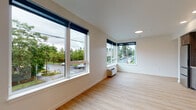 Located on NE Dekum Street in Portland, Oregon, this single-floor unit offers a clean, contemporary design with an open and functional layout. Light hardwood flooring flows throughout the space, creating a warm, cohesive aesthetic that ties each area together seamlessly.

The main living area is bright and airy, featuring large corner windows fitted with roller shades that frame views of the surrounding tree-lined neighborhood. A wall-mounted HVAC unit provides efficient climate control. The open sightlines between the living space and kitchen make the layout feel expansive and connected.

The kitchen is well-appointed with rich dark wood cabinetry extending to the ceiling, providing ample storage. A full suite of stainless steel appliances includes a refrigerator, electric range, over-the-range microwave, and dishwasher, all set against clean, light countertops and a simple, modern backsplash.

A long, recessed-lit hallway connects the living areas to the private spaces, maintaining a tidy and organized flow through the unit. The bathroom is finished in a clean, neutral palette featuring a full-size soaking tub with shower, chrome fixtures, and an accessibility grab bar, all accented by the same light hardwood-style flooring found throughout.

Additional windows in the bedroom offer continued views of lush greenery, keeping the space feeling bright and connected to the outdoors.