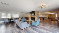 This generously sized single-floor community clubhouse at Village Green offers a thoughtfully designed open layout that accommodates a variety of social and recreational activities. The welcoming entrance features a rustic sliding barn door, patterned carpet, and a pair of accent chairs that set an inviting tone. The main lounge area is anchored by a striking floor-to-ceiling stacked stone fireplace with a wall-mounted television, complemented by a seating arrangement of contemporary sofas and accent chairs atop patterned area rugs. Wide-plank hardwood-style flooring flows throughout, tying the space together cohesively. The fully equipped kitchen includes stainless steel appliances, dark cabinetry with a tile mosaic backsplash, a large island with a light stone countertop, and stylish bar stools beneath pendant lighting — with large windows offering outdoor views. Adjacent dining areas feature multiple round tables with upholstered chairs, bathed in natural light from white-trimmed windows with horizontal blinds. A dedicated games area includes a pool table, shuffleboard table, and exposed brick accent wall, while a separate room provides ample space for ping pong. Throughout, two-tone walls, flush-mount lighting, and consistent flooring create a polished, welcoming atmosphere.