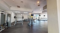 This spacious fitness center is thoughtfully designed to accommodate a full range of workout needs across a single open floor. The layout is divided into distinct functional zones, each optimized for a specific type of training. The cardio area is expansive and bright, featuring a row of treadmills and elliptical machines positioned alongside floor-to-ceiling windows and glass entry doors that invite natural light and outdoor views of surrounding greenery. Ceiling fans overhead enhance air circulation throughout this section. The strength training zone is well-appointed with a power rack, a red-accented squat rack, cable machines, a dumbbell rack, and flat benches, all positioned on durable rubber flooring designed to handle heavy equipment. A functional training area with open floor space, wall-mounted televisions, and teal accent walls provides room for bodyweight and dynamic exercises. Full-length mirrors line sections of the walls, enhancing the sense of space and allowing users to monitor their form. Structural columns painted in teal and yellow add a modern, energetic aesthetic. Recessed and fluorescent lighting supplements the natural light throughout. The overall ambiance is clean, modern, and motivating, with a neutral white and gray palette accented by bold color choices that give the space a contemporary fitness facility feel.