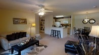 This single-floor residence offers an open, airy layout that seamlessly connects the living area to the kitchen, creating a cohesive and functional living environment. The living room is generously sized, featuring soft neutral carpeting, a ceiling fan with integrated lighting, and built-in shelving along one wall — ideal for displaying décor and personal items. A framed piece of wall art adds a finishing touch to the warm, inviting ambiance.

The kitchen is positioned at the heart of the home, separated from the living area by a peninsula-style breakfast bar with seating for multiple stools. White cabinetry lines the kitchen walls, complemented by standard appliances including a refrigerator and microwave. Under-cabinet lighting adds a practical and welcoming glow to the space.

Natural flow between the living and dining areas encourages easy entertaining and everyday comfort. The consistent neutral color palette — from the walls to the carpeting — creates a calm, cohesive atmosphere throughout. A dedicated dining area with additional seating is visible adjacent to the kitchen. The layout is thoughtfully designed to maximize usable space while maintaining an open feel, making this a comfortable and practical single-level home.
