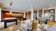 This single-floor amenity space is thoughtfully designed as a multifunctional social hub, blending entertainment, relaxation, and casual gathering areas within one cohesive layout. The interior design embraces a contemporary aesthetic, combining warm earth tones, bold accent walls, and rich mixed-material finishes to create an inviting yet energetic atmosphere.

The game room area is a central highlight, featuring a full-size billiards table positioned beneath modern linear pendant lighting. Additional entertainment options include a foosball table and a classic arcade game cabinet, while a wall-mounted flat-screen TV adds to the entertainment offerings. A seating area with plush, patterned lounge chairs and textured area rugs provides a comfortable spot for casual conversation or spectating.

The bar and lounge zone is anchored by a sleek white quartz waterfall island with bar-height seating, complemented by striking black-and-gold drum pendant lights overhead. Dark wood cabinetry, under-counter refrigeration, and a dramatic floor-to-ceiling abstract wallcovering in earthy bronze and slate tones serve as a bold focal point behind the bar. Light wood-toned tile flooring transitions seamlessly throughout this area. Large windows and glass doors connect to an outdoor terrace, allowing natural light to fill the space.