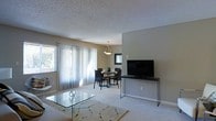 This single-floor space features an open-concept layout that seamlessly connects the living and dining areas, creating a cohesive and functional environment. The living area is anchored by plush wall-to-wall carpeting, a glass-top coffee table, and a patterned area rug, contributing to a clean and understated aesthetic. A sleek media console with a large flat-screen television is positioned along one wall, providing a clear focal point for the room.

The dining area transitions naturally from the living space and is illuminated by a pendant light fixture that hangs above a compact dining table surrounded by dark-framed chairs. A framed mirror on the adjacent wall adds depth to the space.

Large sliding glass doors dressed in sheer white vertical blinds dominate one wall, allowing abundant natural light to filter into the interior while offering a visual connection to the outdoors. A side window further enhances the brightness of the room. The textured popcorn ceiling and neutral warm-toned walls provide a simple backdrop that ties the entire space together, lending it a welcoming and practical character.