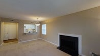 Located at 100 Hempstead Ln in Cary, North Carolina, this single-floor residence offers a comfortable and functional layout designed for everyday living. The main living area is a welcoming space anchored by a classic fireplace with a clean white surround and dark firebox, adding both warmth and a focal point to the room. Neutral-toned walls and wall-to-wall carpeting create a soft, cohesive atmosphere throughout, while a window allows natural light to brighten the space. A stylish pendant light fixture hangs overhead, providing additional illumination and a decorative touch. The living area flows seamlessly into the kitchen via a convenient pass-through opening, enhancing the open feel of the floor plan. The kitchen features white cabinetry, standard appliances including a refrigerator and range, and a countertop ledge that doubles as a casual dining or serving area. Crisp white trim and baseboards frame the rooms, adding a polished, finished look throughout. The single-level design ensures easy accessibility and a straightforward flow between spaces, making this home practical and inviting.