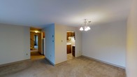 This two-story residence offers a functional and comfortable layout spread across two levels. The main living area features wall-to-wall carpeting that adds warmth underfoot, while neutral-toned walls create a clean, versatile backdrop throughout. An elegant chandelier with classic tulip-style shades serves as a focal point in the dining area, providing both ambient and task lighting. Adjacent to the dining space, the kitchen is efficiently arranged with rich, warm-toned wood cabinetry providing ample storage, and tile flooring that contrasts neatly with the carpeted living zones. The kitchen layout includes upper and lower cabinets, offering generous storage and counter space. A hallway connects the main living areas to the staircase, which leads to the second floor, and a full-size refrigerator is visible along the corridor. The staircase, finished with carpeted treads and a handrail, provides access to the upper level. The overall design emphasizes practicality with a straightforward floor plan, making good use of available space. Natural and artificial lighting work together to keep the interior feeling open and well-illuminated throughout both floors.