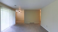 This single-floor space presents an open living area finished with wall-to-wall carpeting in a neutral tone, creating a cohesive and understated aesthetic throughout. The layout is defined by a partial dividing wall that separates the main living zone from a secondary area beyond, offering a sense of distinct spaces while maintaining an open feel. A ceiling fan with an integrated light fixture is mounted in the secondary area, providing both airflow and ambient lighting. The textured popcorn ceiling adds a classic touch, while flush-mounted HVAC vents in both the ceiling and floor ensure climate control throughout. Neutral beige walls complement the carpeting and contribute to a warm, understated atmosphere. On one side of the main living area, vertical blinds frame what appears to be a sliding glass door, allowing natural light to filter in. A hallway visible in the background suggests additional rooms beyond the main living space. The overall layout is straightforward and functional, offering a blank canvas well-suited for a variety of furniture arrangements and décor styles.