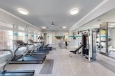 Located at 24215 Kuykendahl Road in Tomball, Texas, this fitness center offers a spacious, single-floor layout designed to accommodate a comprehensive range of workout activities. The interior presents a clean, modern aesthetic defined by light gray walls accented with a bold teal horizontal stripe that runs throughout the space, complemented by light wood-style flooring that enhances the bright and open atmosphere.

The main exercise area is generously sized to house an impressive selection of cardio equipment, including multiple treadmills, elliptical trainers, a stair climber, and a rowing machine. A dedicated strength-training section features a multi-station cable machine, a weight bench, a full dumbbell rack, and kettlebells of varying sizes, along with a battle rope for high-intensity training. Floor-to-ceiling mirrors line key sections of the walls, visually expanding the space and allowing users to monitor their form during workouts.

Natural light filters in through several windows positioned near the entry and along the walls, while flush-mount ceiling lights and ceiling fans ensure consistent illumination and air circulation. A wall-mounted flat-screen television adds an entertainment element to the space. Decorative framed artwork and a sculptural wall piece on the teal accent wall contribute a polished, welcoming ambiance. The facility is accessible via a dedicated entrance with an exterior door and transom window.