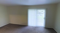 This two-floor space presents a straightforward and functional design. The living area, visible in the provided image, features wall-to-wall carpeting in a neutral tone, contributing to a comfortable and understated ambiance. A full-width sliding glass door serves as a prominent architectural feature, fitted with vertical blinds that allow for adjustable light control. The door provides a direct view of mature trees and greenery outside, bringing a natural element into the interior and allowing ample natural light to fill the room. The walls are finished in a soft, warm neutral tone, complementing the light carpeting and creating a cohesive, calm atmosphere. Smooth white ceilings complete the clean aesthetic of the space. Spanning two floors, the layout offers separation between living areas, providing versatility for various uses across levels. The overall design is practical and well-suited for everyday living, with an emphasis on natural light and connection to the outdoors through the sliding glass door access point.