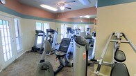 This single-floor fitness center is designed to support a well-rounded workout experience. The space is outfitted with a comprehensive selection of cardio equipment, including treadmills, elliptical trainers, stationary bikes, and stair-climbing machines — all arranged to maximize usability within the room. Full-length mirrored walls line one side of the space, creating a sense of openness and allowing users to monitor their form during exercise. The room features a warm color palette with terracotta-toned upper walls and a sage green accent band, giving the space an inviting, approachable ambiance. A ceiling fan is centrally mounted overhead, providing consistent air circulation during workouts. Multiple windows along the perimeter allow natural light to filter in, brightening the interior and offering a connection to the outdoors. The textured ceiling and patterned carpet flooring add a comfortable, community-style character to the space. Overall, this fitness center provides a practical and comfortable environment suitable for a variety of cardiovascular training routines.