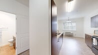 This single-floor residence offers a well-organized layout with distinct living, sleeping, and utility areas. The kitchen is a standout feature, equipped with rich dark-brown cabinetry extending to the ceiling, light countertops, black appliances including a range, microwave, and refrigerator, and tile flooring in a neutral stone tone. A peninsula counter with additional storage separates the kitchen from the adjacent dining area, which benefits from natural light through a window and is accented by a multi-bulb chandelier overhead. A wall-mounted HVAC unit provides climate control in the dining area.

The bedrooms are carpeted in a warm beige tone, offering a comfortable feel underfoot. One bedroom features vaulted ceilings and a large window with outdoor views, adding a sense of openness. Additional bedrooms are equally bright, each with windows allowing natural light and views of the surrounding neighborhood.

The bathroom includes tile flooring, a walk-in glass-enclosed shower, a toilet, and a single vanity with dark wood cabinetry and a light countertop. Storage solutions are present throughout, including built-in shelving in what appears to be a closet area. The overall palette of whites and neutrals creates a clean, cohesive aesthetic.