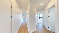 This contemporary single-floor residence showcases a clean, modern design with a thoughtfully planned layout. Light-toned hardwood flooring flows throughout the main living areas and hallways, lending a cohesive and airy feel to the space. A long, well-lit corridor connects the various rooms, lined with flush panel doors featuring matte black hardware that adds a refined, minimalist touch.

The open-concept living and kitchen area is a standout feature, offering generous space for both relaxation and entertaining. The kitchen is equipped with stainless steel appliances—including a full-size refrigerator, range, and microwave—complemented by warm wood cabinetry and white upper cabinets. A large window in the kitchen allows abundant natural light to pour in.

Bedrooms are carpeted for comfort, each equipped with a wall-mounted mini-split HVAC unit, and one features a floor-to-ceiling window with double doors that open to an outdoor view, filling the room with natural light.

The primary bathroom is elegantly appointed with large-format tile flooring, a glass-enclosed walk-in shower with tile surround, a sleek floating wood vanity, and a backlit mirror. A separate half bath provides added convenience with matching wood cabinetry and tile flooring. The overall ambiance is bright, clean, and modern throughout.