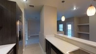 This two-story residence showcases a thoughtfully designed interior with clean, contemporary finishes throughout. The kitchen is a standout feature, highlighted by an expansive quartz countertop with subtle speckled detailing, complemented by dark cabinetry with sleek hardware. A pendant light with a warm amber glass shade hangs above, adding a refined focal point to the space.

The open layout connects the kitchen to adjoining living areas, with a hallway leading to additional rooms on the main level. Light-toned tile flooring runs throughout, contributing to a bright and cohesive aesthetic. The neutral wall tones in soft gray enhance the sense of openness and complement the overall color palette.

A doorway visible from the kitchen area provides access to an outdoor balcony, allowing natural light to filter into the space through a slatted window blind. The second floor adds additional living space, with the staircase positioned conveniently along the main corridor. Recessed lighting and ceiling-mounted vents are integrated cleanly into the design, ensuring both comfort and a polished appearance throughout the home.