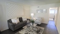 This well-appointed single-floor space offers a thoughtfully designed interior with a clean, modern aesthetic. The living area serves as the centerpiece of the space, showcasing rich dark hardwood floors that contrast beautifully with the bright, neutral-toned walls, creating a warm yet contemporary atmosphere.

The living room is furnished with a plush charcoal gray tufted sofa accented by an assortment of decorative pillows, complemented by a patterned accent chair that adds visual interest and texture. A sleek glass and chrome coffee table anchors the seating arrangement, resting atop a layered area rug that adds depth and coziness to the space. Decorative elements, including a large geometric wall art piece and an abstract canvas, add personality and dimension to the walls.

Ambient lighting is provided by a classic table lamp, enhancing the inviting atmosphere of the living area. Visible ceiling vents suggest a central HVAC system for year-round comfort. The overall design palette of grays, whites, and silvers creates a cohesive, sophisticated look throughout. The space is styled with careful attention to detail, balancing functionality and aesthetics to deliver a comfortable and visually appealing environment.
