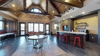 The Retreat is a thoughtfully designed single-floor communal space that seamlessly blends rustic charm with modern sophistication. The most striking architectural feature is the dramatic vaulted ceiling clad in rich wood paneling, accented by exposed dark timber beams that draw the eye upward and lend the space a grand, lodge-inspired character. Arched upper windows allow natural light to pour in, enhancing the warm, inviting atmosphere throughout.

The open-plan layout is divided into distinct yet connected functional zones. A lounge area near the large windows offers comfortable seating with red accent cushions, ideal for relaxed gatherings. At the center of the space, a round dining table with mixed seating provides a casual spot for socializing. The floors feature a striking herringbone-patterned tile that transitions to a darker slate-style finish near the bar area, adding visual depth and texture.

The full-length bar counter, finished in crisp white, is a centerpiece of the space, lined with vibrant red metal bar stools that provide a bold pop of color. Behind the bar, a stone accent wall frames a mounted television, flanked by industrial-style gooseneck lighting and open shelving. The overall ambiance is energetic yet comfortable, making The Retreat an ideal venue for community events and everyday gatherings alike.