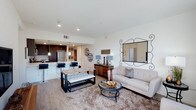 This single-floor residence showcases an open-concept layout that connects the living area and kitchen into one cohesive, well-lit space. The living area is anchored by a plush area rug and features a neutral-toned sofa paired with a round glass-top coffee table, creating a comfortable yet contemporary seating arrangement. A tufted bench and rustic console table add layered texture and warmth to the space. A decorative wrought-iron mirror serves as a prominent wall accent, enhancing the sense of depth and visual interest.

The kitchen flows naturally from the living space and is equipped with stainless steel appliances, dark cabinetry, and a light countertop breakfast bar with seating for multiple guests. Pendant lighting above the bar adds a refined touch, complementing the recessed lighting throughout the space. The flooring transitions seamlessly between wood-look planks in the kitchen and dining areas and plush carpeting in the living zone. Bright white walls and an open layout allow natural and artificial light to circulate freely, contributing to an airy, welcoming atmosphere throughout.