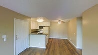 This single-floor residence offers a well-designed open-concept layout that seamlessly connects the kitchen and living area. Rich, warm-toned hardwood flooring runs throughout the main living space, adding a sense of warmth and cohesion to the interior.

The kitchen is positioned as a functional centerpiece, featuring a peninsula with a dark countertop that provides additional prep space and acts as a natural divider between the cooking and living zones. White cabinetry lines the upper walls, offering ample storage, while appliances include a built-in microwave, an electric range, and a full-size refrigerator. A recessed ceiling light fixture directly above the kitchen ensures well-lit meal preparation.

The adjacent living area is open and airy, finished with the same hardwood flooring and complemented by a ceiling fan with an integrated light fixture — a practical feature that aids in air circulation and ambient lighting. Neutral warm-beige walls provide a versatile backdrop throughout. White baseboards and trim add a clean, finished look to the space.

Textured ceilings are present throughout, adding a subtle architectural detail. The layout is efficient and functional, making strong use of the available floor space to create a comfortable, connected living environment.