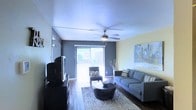 This two-floor residential space offers a well-designed interior that balances comfort and modern style. The main living area immediately draws attention with its rich hardwood flooring, which adds warmth and character throughout the space. A ceiling fan centered overhead provides both functionality and a polished finishing touch, while a textured ceiling adds subtle visual interest above.

The living room is generously proportioned, featuring a contemporary layout with a plush gray sofa accented with decorative pillows, a tufted accent chair, and a round, low-profile coffee table anchoring the seating arrangement atop a patterned area rug. A large flat-screen television is positioned on a dark media console along one wall, creating a dedicated entertainment zone.

Two-toned walls — a bold gray paired with a soft yellow-green — give the space a modern, curated aesthetic. Natural light flows abundantly into the room through a wide sliding glass door fitted with vertical blinds, which opens to an outdoor area and enhances the overall sense of openness. Decorative artwork on the wall adds a personal, artistic touch to the ambiance.

The second floor extends the living space further, offering additional functional areas. Overall, this residence presents a cohesive, contemporary environment with thoughtful design details throughout.