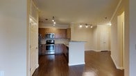 This single-floor residence offers a thoughtfully designed open-concept layout that seamlessly connects the kitchen and living areas. The kitchen is a functional centerpiece, showcasing warm medium-toned wood cabinetry, a classic tile backsplash, and a full suite of stainless steel appliances including a refrigerator, range, and built-in microwave. A peninsula with granite countertops extends the workspace and creates a natural divider between the kitchen and the adjoining living area, making it ideal for both cooking and entertaining. Rich, dark hardwood flooring runs throughout the space, adding warmth and a sense of continuity. Crown molding along the ceiling lends an elevated, polished finish to the interior, while recessed track lighting provides adjustable, well-distributed illumination across both the kitchen and living zones. The open living area is generously proportioned, offering flexible space for a variety of furniture arrangements. Neutral warm-toned walls complement the natural wood tones and create a welcoming, cohesive ambiance. Additional doors visible from the main area suggest access to further rooms, contributing to a well-organized and practical floor plan throughout.