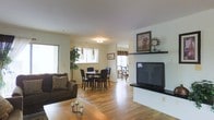 This two-floor space showcases a well-designed open-concept main living area that integrates the living room, dining area, and additional spaces into a cohesive, flowing layout. The living room is anchored by a clean white fireplace mantel, above which decorative artwork and a clock are displayed, adding a touch of character to the space. A large flat-screen television sits on a media console beside the fireplace, making it a functional entertainment focal point. The living room features a comfortable dark upholstered sofa paired with a wooden coffee table, complemented by warm hardwood flooring that extends throughout the main level. The dining area is positioned adjacent to the living room and accommodates a round dark wood dining table surrounded by matching chairs, bathed in natural light streaming through multiple windows with light curtains. Potted indoor plants add a natural element to the space. Beyond the dining area, a further room is visible, suggesting additional living or recreational space. The overall ambiance is warm, bright, and inviting, with neutral wall tones and natural light enhancing the sense of openness across both levels.