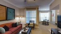 This single-floor residential space offers a thoughtfully designed interior that balances comfort and functionality. The living area serves as the heart of the home, showcasing warm neutral wall tones complemented by plush carpeted flooring that adds a sense of coziness underfoot. Large windows dressed with white horizontal blinds and elegant grommet-top curtains allow generous amounts of natural light to fill the room, creating a bright and airy atmosphere throughout the day. Crown molding along the ceiling adds a refined architectural touch, lending the space a polished, well-appointed feel. A ceiling fan overhead provides both practical airflow and a subtle design element. The living area is well-suited for relaxation and entertaining, with ample space for seating arrangements. Adjacent to the main seating area, a compact dining nook features a bistro-style table and stools, offering an intimate setting for casual meals. The overall ambiance is warm and welcoming, with a cohesive design palette that ties the living and dining areas together seamlessly. The abundance of natural light, classic architectural detailing, and neutral color scheme combine to create a comfortable and visually appealing living environment.
