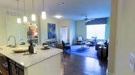 This single-floor residence is designed with an open-concept layout that seamlessly connects the kitchen, dining, and living areas into one cohesive living space. The kitchen features a granite countertop peninsula with pendant lighting overhead, providing both a functional workspace and a casual dining option. Rich hardwood flooring flows throughout the main living area, adding warmth and a sense of continuity to the space. The living room is generously proportioned, anchored by a ceiling fan and framed by a large window dressed with bold geometric-patterned valances that allow natural light to fill the room. A vibrant area rug in blue tones adds a pop of color and defines the seating area. Comfortable, stylish furnishings complement the contemporary aesthetic, while colorful wall art contributes to the lively, welcoming ambiance. A glimpse into an adjacent room reveals additional living or sleeping space, suggesting a thoughtful and functional floor plan. The overall atmosphere is bright, modern, and inviting, making the most of its open layout and natural light.