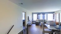 This two-story space showcases a well-designed interior that balances practicality with modern style. The main living area is anchored by rich hardwood flooring that extends throughout, providing warmth and visual continuity. A bold blue accent wall in the living room creates a striking focal point, complementing the neutral white walls found elsewhere in the space.

The living area is generously sized, accommodating a sectional sofa, ottoman, and additional seating, with multiple windows dressed in dark curtain panels that allow for natural light control. The windows invite ample daylight into the room, enhancing its airy feel. A slim metal-framed console table adds a contemporary, industrial touch to the décor.

A staircase with a dark wood banister connects the two floors, visible from the main living area and adding architectural interest. An adjacent room, likely a bedroom, is partially visible through a doorway, suggesting a thoughtful layout that separates living and sleeping areas. The overall ambiance is modern and inviting, with a neutral palette punctuated by carefully chosen design accents throughout.