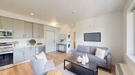 Located at 7460 Southwest Hermoso Way in Tigard, Oregon, this thoughtfully designed 2-bedroom, 1-bathroom residence offers a clean, modern aesthetic throughout its efficiently organized single-floor layout. Light wood flooring flows seamlessly across every room, contributing to a bright and cohesive atmosphere.

The open-concept kitchen and living area serves as the heart of the home. The kitchen features contemporary gray cabinetry extending to the ceiling, white subway tile backsplash, light quartz countertops, and a full suite of stainless steel appliances including a refrigerator, range, over-the-range microwave, and dishwasher. The adjacent living area is well-lit and includes space for both a dining table and a comfortable seating arrangement, with a stacked in-unit washer and dryer conveniently tucked nearby.

The primary bedroom is generously proportioned, bathed in natural light from a wide row of windows overlooking lush, mature greenery. The second bedroom includes a sliding-door closet and ample space. The full bathroom is clean and bright, finished with gray vanity cabinetry, a tub-shower combination, and matching light flooring. Throughout the residence, white walls and recessed lighting enhance the open, airy feel of the space.