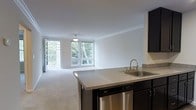 This single-floor unit showcases an open-concept layout that seamlessly connects the kitchen and living area, creating a bright and airy atmosphere. The kitchen is outfitted with dark espresso cabinetry, a stainless steel sink with a brushed nickel faucet, a stainless steel dishwasher, and a granite-style countertop that extends into a peninsula — providing additional prep space and a natural divider between the kitchen and living area. Crown molding runs along the ceiling throughout, adding a refined architectural detail to the otherwise clean, white interior. The living area features neutral wall-to-wall carpeting and is bathed in natural light from large windows and a sliding glass door that opens onto a private balcony. The outdoor views showcase mature trees and lush greenery, lending a serene, nature-connected feel to the interior. A pendant light fixture is centrally positioned over the living space, complementing the natural light. The overall ambiance is clean, modern, and inviting, with a cohesive neutral palette that allows flexibility for various interior design styles.