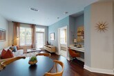 This well-appointed single-floor residence combines contemporary design with functional living across its thoughtfully arranged layout. The kitchen serves as a visual centerpiece, showcasing a striking two-tone cabinetry scheme of bold navy lower cabinets paired with crisp white uppers, accented by gold hardware and pendant lighting. Stainless steel appliances, a herringbone tile backsplash, white quartz countertops, and a waterfall-edge island with a brass-finish sink and built-in seating complete the space. Rich hardwood flooring flows seamlessly from the kitchen into the open living area, where large windows admit generous natural light and provide views of surrounding greenery. A glass-paneled door extends the living room toward an outdoor space. A built-in desk nook integrated into the living area offers a dedicated workspace. The bedroom features warm, carpeted flooring, an eye-catching terracotta accent wall, and ample natural light from large windows. The en-suite bathroom continues the navy cabinetry theme with a wide double vanity, white quartz countertop, and dark hardwood flooring. Throughout, the residence balances bold color choices with clean, modern finishes to create a cohesive and inviting atmosphere.