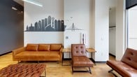 Suite D66 is a thoughtfully designed commercial office space that balances privacy with an open, collaborative atmosphere. The suite is accessed through a bright, glass-fronted entry featuring floor-to-ceiling windows and double glass doors that flood the reception area with natural light and offer views of the surrounding urban landscape. Warm hardwood-style flooring runs throughout the space, lending a cohesive and professional aesthetic.

A central corridor connects the suite's various private offices and meeting rooms, all enclosed by sleek black-framed glass partition walls. This design maintains transparency and openness while providing acoustic separation for focused work. The private offices are furnished with long work surfaces and ergonomic seating, while dedicated conference rooms offer wooden tables and presentation screens suited for team meetings or client sessions.

Common lounge areas are furnished with rich cognac leather sofas, accent chairs, and low tables, creating inviting spaces for informal collaboration. A striking Dallas city skyline mural adorns one wall, adding a strong local character and visual focal point to the communal area. Overhead, suspended linear lighting and track fixtures illuminate the space efficiently. The suite's modern industrial design, glass-forward architecture, and carefully curated furnishings make it a functional and professional environment well-suited for dynamic business operations.