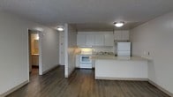 This single-floor space showcases an open-concept layout that seamlessly connects the kitchen and living areas, creating a functional and cohesive environment. The kitchen is equipped with white cabinetry, offering ample upper storage space, and includes a full suite of white appliances — a refrigerator, electric range, and built-in range hood. A double sink is positioned along the counter, complemented by a light-toned countertop that extends to form a peninsula, providing additional workspace or casual dining potential. Dark hardwood-style flooring runs throughout the main area, offering a strong contrast against the white kitchen finishes and neutral grey walls. Flush-mount ceiling light fixtures illuminate the space evenly, while a textured ceiling adds subtle character. A structural column near the kitchen helps define the layout without fully closing off the space. A hallway visible in the background suggests access to additional rooms, and a door provides entry or access to adjacent spaces. Overall, the unit presents a clean, practical interior suited for everyday living.
