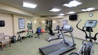 This fitness center offers a practical and functional workout environment designed to support a range of exercise routines. The space is illuminated by a series of recessed skylight-style ceiling panels that provide bright, even lighting throughout the room, complemented by warm wall-mounted accent lights. The flooring is covered in durable, low-pile carpeting suited for a high-traffic exercise environment. A large wall mirror spans one side of the room, visually expanding the space and allowing users to monitor their form during workouts. The equipment layout is thoughtfully arranged, featuring True-brand treadmills and a recumbent stationary bike as the primary cardio offerings. A free weights rack lines the mirrored wall, accompanied by a weight bench, providing options for strength training. Exercise balls in multiple sizes add versatility for stretching and core workouts. A wall-mounted flat-screen television is positioned for viewing during cardio sessions. Informational fitness posters are displayed on the walls, offering workout guidance. The warm, neutral-toned walls contribute to a clean and welcoming atmosphere, making this fitness center a well-rounded facility for both cardiovascular and strength-based training.